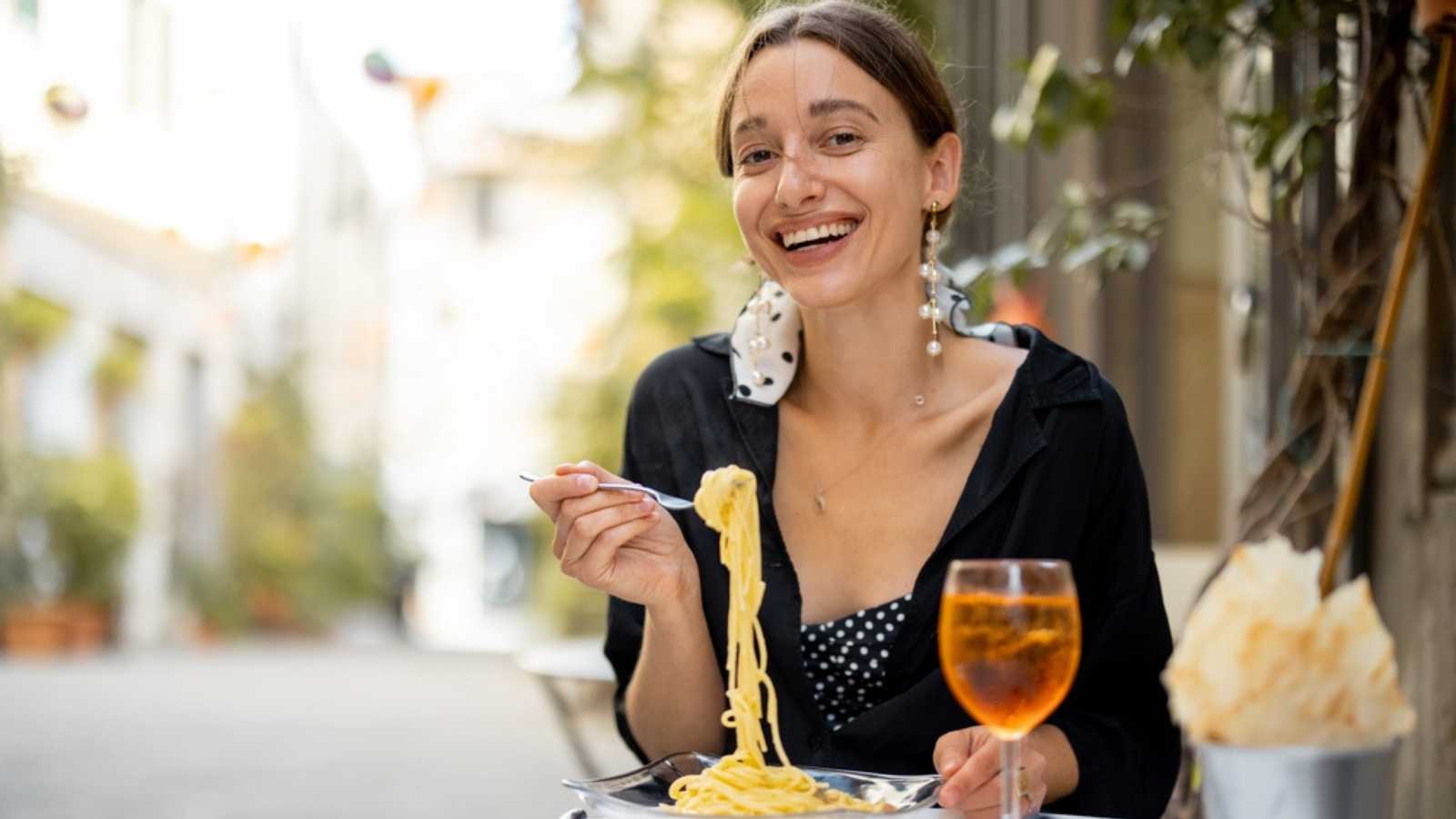 Portrait of a smiling woman eating pasta and wine at cozy italian restaurant outdoors. Concept of italian cuisine and lifestyle. Italian pasta carbonara and Spritz Aperol